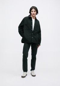 Camisa casaco oversized preta com botões de pressão e dois bolsos frontais, combinada com jeans escuros e ténis brancos. T-shirt branca visível.