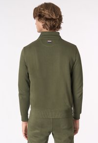 Felpa verde oliva con colletto alto, polsini e orlo a costine. Presenta una piccola patch con logo sul collo. Tessuto dalla texture liscia.