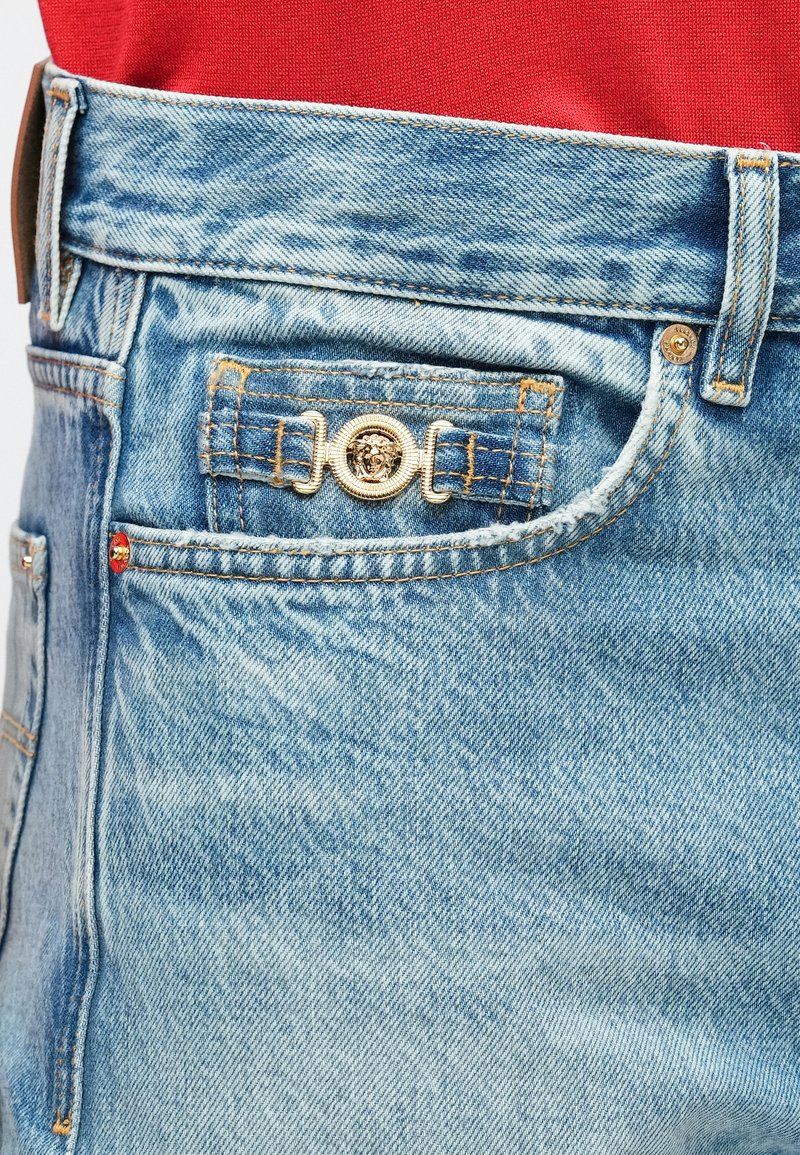 Close-up van een blauw denim jeanszak met een gouden sierknop en rood stof zichtbaar boven de tailleband.