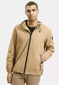 Lekka beżowa kurtka z zamkiem z przodu, regulowanym kapturem, elastycznymi mankietami i bocznymi kieszeniami, wykonana z tkaniny wodoodpornej.