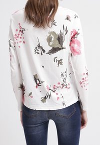 Långärmad vit topp med rosa och gröna blommönster. Mjuk tyg med rundad nederkant och sidoflikar, i kombination med mörka denimjeans.