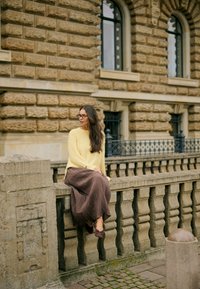 Femme aux cheveux longs et lunettes, portant un pull jaune et une jupe marron, assise sur une balustrade en pierre devant un bâtiment historique.