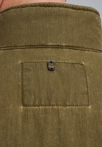 Chemise à manches courtes de couleur olive avec une texture lisse, ornée d'un petit emblème noir et d'une poche en patch au dos.