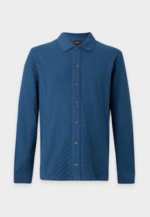 Man staat en draagt een blauw gestructureerd overhemd met knopen, een wit ondershirt, lichtblauwe wijde jeans en donkerbruine schoenen tegen een effen achtergrond.