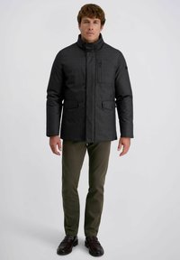 Schwarze gesteppte Jacke mit hohem Kragen, vorderem Reißverschluss und zwei Seitentaschen. Getragen über olivfarbenen Hosen und dunklen Schuhen.