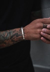 Hõbedane metallist mansettkät tõmblusega viimistlusega, kantud tätoveeritud küünarvarrel. Must lühikeste varrukatega särk on osaliselt nähtav.
