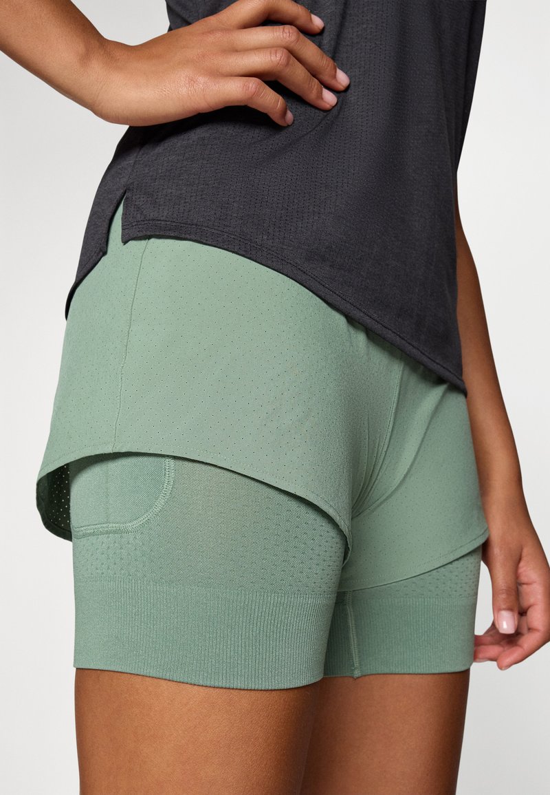 Pantalones cortos deportivos verde claro con una textura perforada, diseño en capas y una cinturilla acanalada, combinados con una parte superior gris oscura.