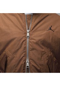 Giacca bomber marrone con chiusura a zip, dotata di una cerniera argentata e logo nero. Tessuto texturizzato e tasche laterali visibili.