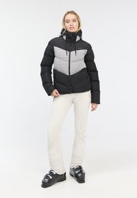 Black and grey puffer jacket with a chevron pattern, featuring a zip closure and hood. Model wears beige pants and black ski boots.