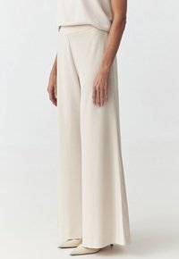 Pantalons beige à jambes larges en tissu lisse, dotés d'une taille mi-haute et d'une silhouette fluide, associés à des talons aiguilles beiges.