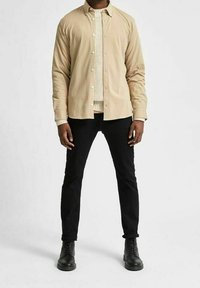 Ljus beige knapp-up skjorta med en slät textur över en krämfärgad stickad tröja, kombinerad med svarta slim-fit byxor och svarta snörstövlar.