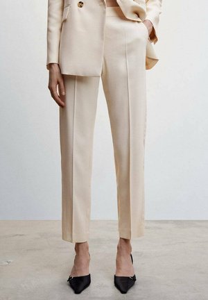 Beige op maat gemaakte broek gecombineerd met een bijpassend colbert, gedragen met zwarte pumps met spitse neus op een neutrale vloerachtergrond.