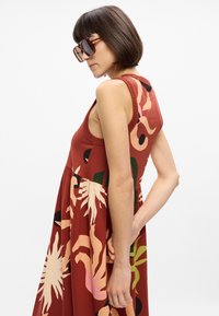 Šaty bez rukávů s květinovým vzorem v rezavě oranžové barvě, s nařaseným pasem. Obsahují výrazné akcenty v růžové, zelené a černé barvě. Vyrobené z lehké tkaniny.
