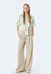 Bloemige shirt met korte mouwen in het wit met groene en beige patronen, geknoopt in de taille; gecombineerd met losse beige broek en gevlochten instappers.