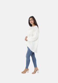 Witte oversized gebreide trui met een ronde hals en ballonsleeves, gecombineerd met blauwe skinny jeans en doorzichtige sandalen met hakken.
