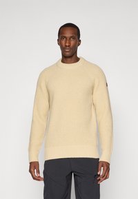Pull en maille crème avec une surface texturée, col rond côtelé et poignets côtelés, porté sur un pantalon sombre. Détail de logo marron sur la manche.