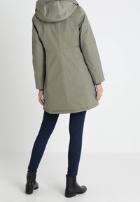 Olivově zelená parka bunda s kapucí, s rovný střihem, zapínáním na zip a hladkou texturou. Kombinována s tmavě modrými úzkými džíny a černými botami.