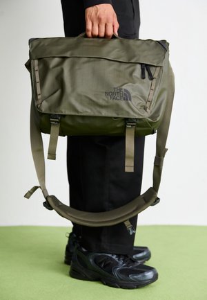 Personne en pantalon et chaussures noirs tenant un sac messager The North Face vert olive par sa poignée sur un fond uni.
