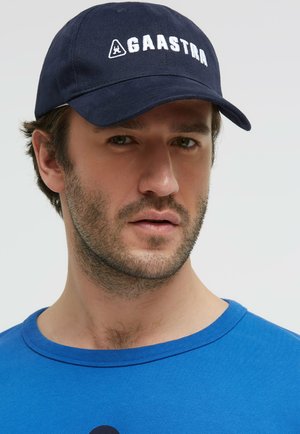 Mann mit kurzem Bart, der eine marineblaue Kappe mit dem Logo "GAASTRA" und ein blaues Hemd vor einem einfarbigen hellen Hintergrund trägt.