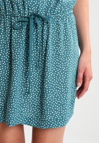 Turquoise jurk met witte stippen, ingesnoerde taille en een stoffen riem die is gestrikt. Lichtgewicht, gestructureerd materiaal, ontwerp tot op de knie.