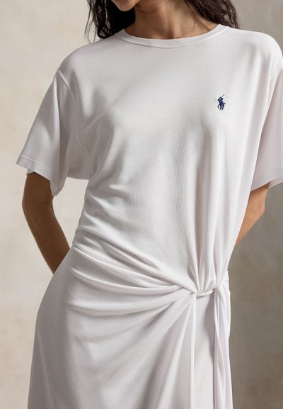 Γυναίκα που φοράει λευκό φόρεμα με κοντά μανίκια, με λεπτομέρεια κόμπου στη μέση και μικρό λογότυπο παίκτη πόλο σε ναυτικό μπλε στο στήθος.