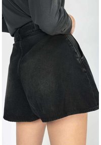 Osoba v černých denimových šortkách s vysokým pasem a postranními kapsami, s tmavým topem s dlouhým rukávem zastrčeným dovnitř, na jednoduchém světlém pozadí.