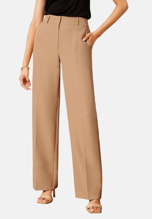 Kvinde iført beige bukser med høj talje og vide ben med sidelommer, kombineret med tanfarvede åbne hæle og en sort ærmeløs top.