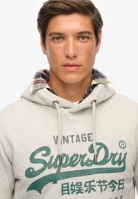 Sudadera gris con logo verde impreso, cordones y detalle de cuello a cuadros, que ofrece un diseño casual pero funcional.