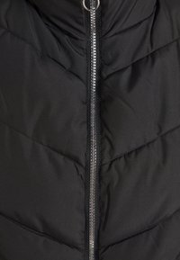Schwarze Steppweste mit strukturiertem Oberflächen, ausgestattet mit einem zentralen silbernen Reißverschluss und einem Zickzack-Steppdesign. Keine auffälligen Muster oder Akzente.