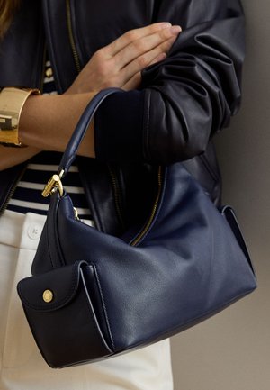 Žena u crnoj kožnatoj jakni i zlatnoj manžetnoj narukvici drži tamnoplavu kožnatu torbu s zlatnim detaljima i patentnim zatvaračem.