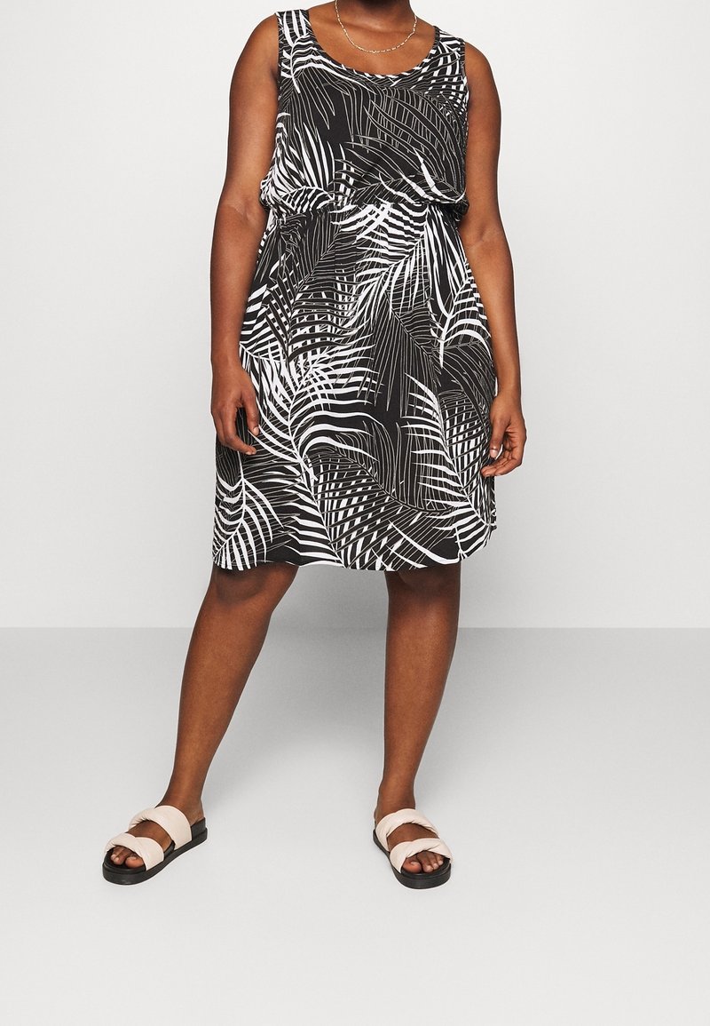 Femme portant une robe noire sans manches avec des motifs de feuilles blanches et des sandales beiges, debout devant un fond clair uni.