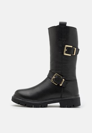 Botte en cuir noir mi-mollet avec deux brides à boucles dorées et une semelle épaisse à crampons, vue de côté extérieur sur un fond uni.