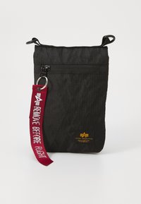 Borsa in nylon nera con superficie testurizzata e zip superiore. Presenta una tracolla rossa con l'etichetta "RIMUOVERE PRIMA DEL VOLO" e un attacco per portachiavi.