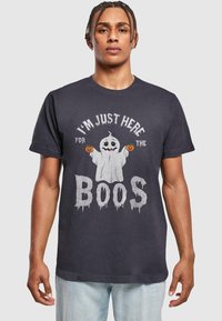 Svart bomullströja med en spökgrafik och pumpor, text som säger "JAG ÄR BARA HÄR FÖR ATT DRICKA", och sliten stil på bokstäverna.