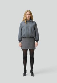 Una donna con capelli ondulati è in piedi contro uno sfondo grigio chiaro, indossando una giacca bomber grigia, una gonna a quadri, collant neri e scarpe a punta.