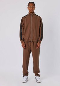 Donkerbruine trainingsoutfit met een ritsjack en bijpassende broek. Bevat zwarte strepen met gemusterde accenten op de mouwen en aan de zijkant van de broek. Witte sneakers.