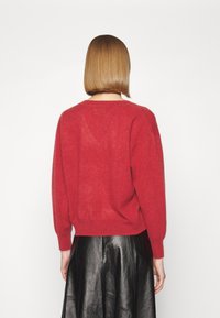 Person med kort, rakt hår som bär en röd långärmad tröja och en svart läderkjol, vänd bort från kameran mot en enkel vit bakgrund.