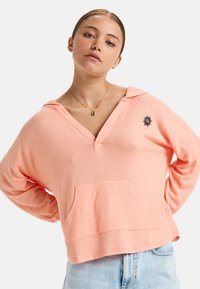 Lichtroze, cropped hoodie gemaakt van zacht materiaal, met een grote voorzak, een v-hals en een geborduurd zonontwerp op de borst.