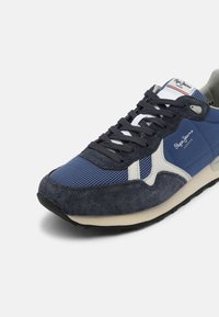 Modrá teniska s kombinací materiálů zířkového a semišového povrchu, s bílými akcenty a černou gumovou podrážkou. Viditelné tkaničky a detail loga.