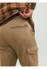Pantaloni cargo beige con una texture liscia, caratterizzati da una tasca posteriore e cuciture decorative. Sovrapposti da una camicia a quadri marrone.