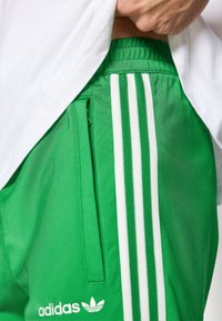 Πράσινο αθλητικό παντελόνι κατασκευασμένο από ελαφρύ υλικό, με λευκές τρεις ρίγες και πλαϊνή τσέπη με φερμουάρ.