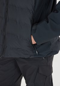 Sort isoleret jakke med kviltet tekstur, frontlynlås og sidelommer. Sammen med sorte bukser med cargo-lommer.