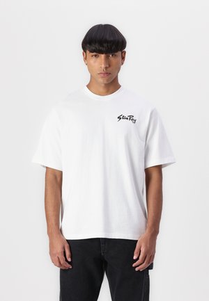 T-shirt bianca in cotone a maniche corte con logo nero sul lato sinistro del petto, scollo rotondo e vestibilità rilassata. Abbinato a jeans scuri.