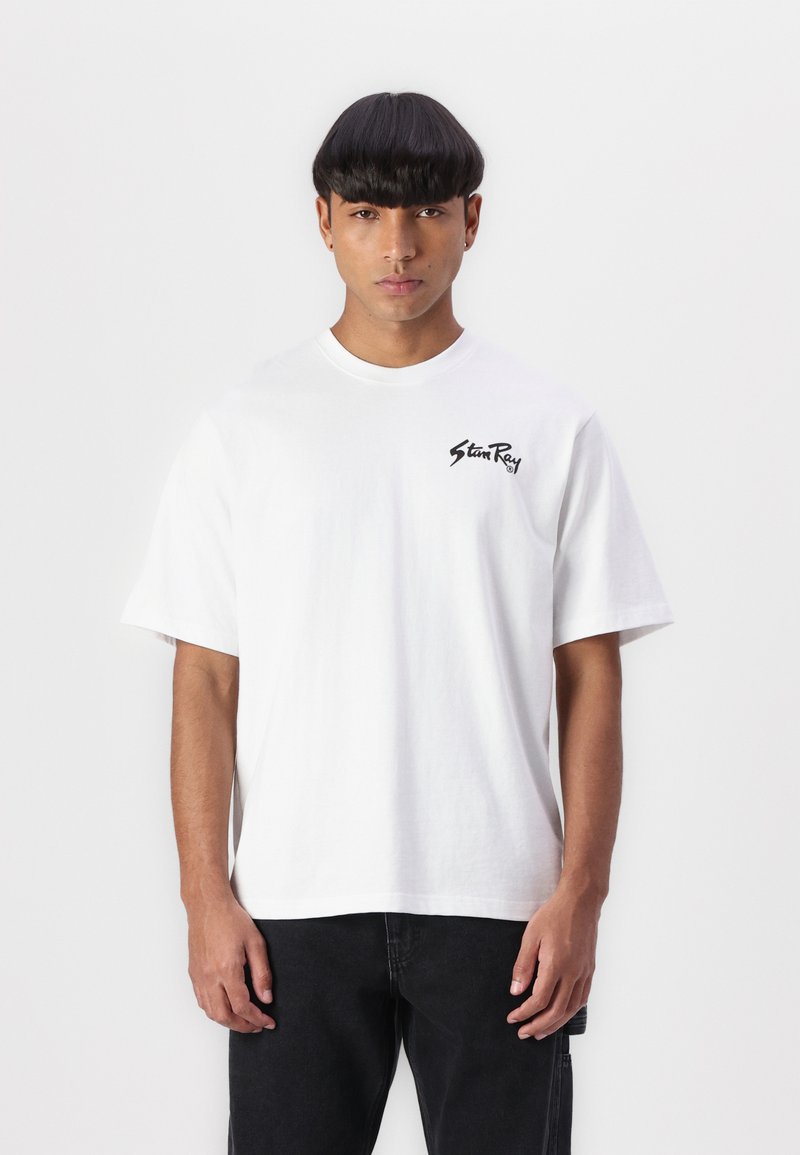 Short-sleeve white cotton t-shirt featuring a black logo on the left chest, crew neckline, and relaxed fit. Paired with dark jeans.