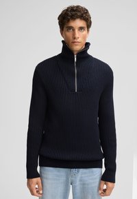 Maglione lavorato a maglia blu navy con colletto alto e chiusura a zip a metà. Presenta un tessuto a trama e orli e polsini a costine. Indossato sopra jeans azzurri.