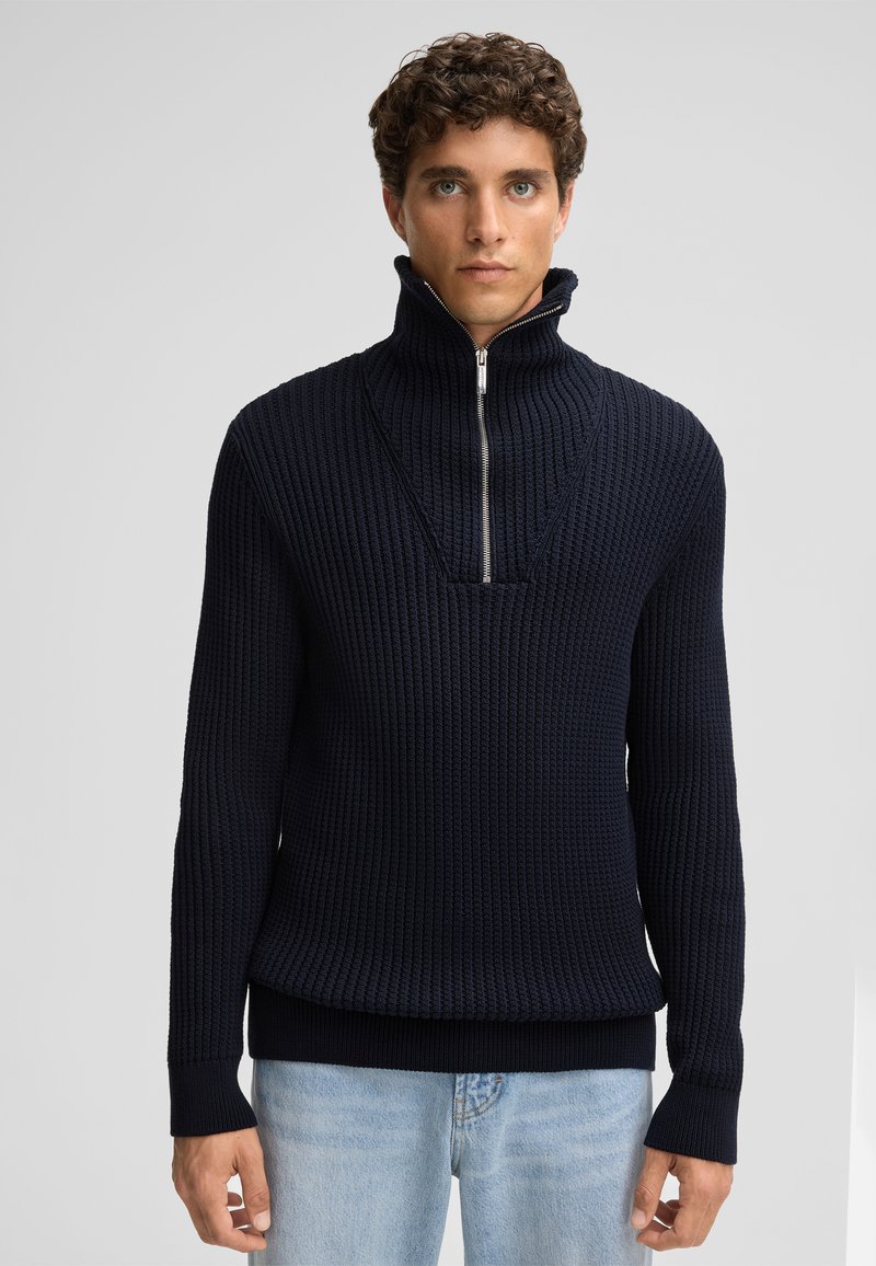 Maglione lavorato a maglia blu navy con colletto alto e chiusura a zip a metà. Presenta un tessuto a trama e orli e polsini a costine. Indossato sopra jeans azzurri.