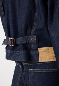 Mørkeblå denimjakke med oransje søm, belte i midjen og et brunt lærmerkelappen med "TOMMY JEANS."