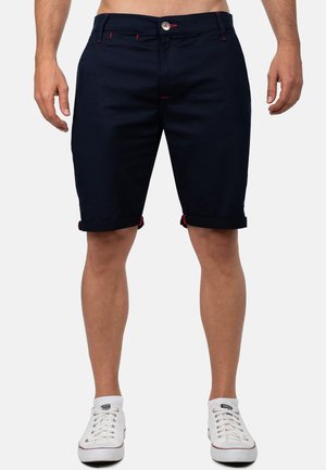 Mann trägt dunkelblaue Shorts mit umgeschlagenem Saum und roten Ziernähten sowie weiße Sneakers mit niedrigem Schaft vor einem einfarbigen Hintergrund.