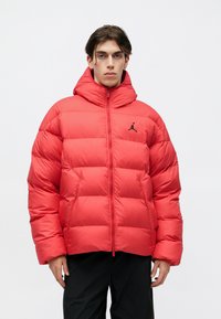 Chaqueta de plumas roja con un cuello alto, cremallera completa y logo negro en el pecho. Presenta secciones acolchadas y capucha. Textura lisa y brillante.