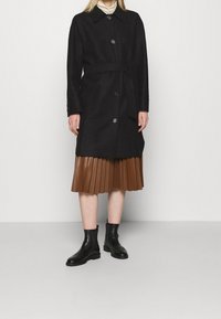 Lungo cappotto nero con colletto, bottoni monopetto e cintura annodata. Abbinato a una gonna plissettata marrone e stivaletti neri alla caviglia.